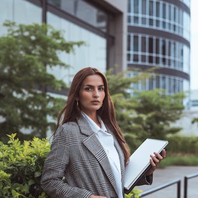 Confident woman in a stylish coat standing outside an office building holding a laptop.