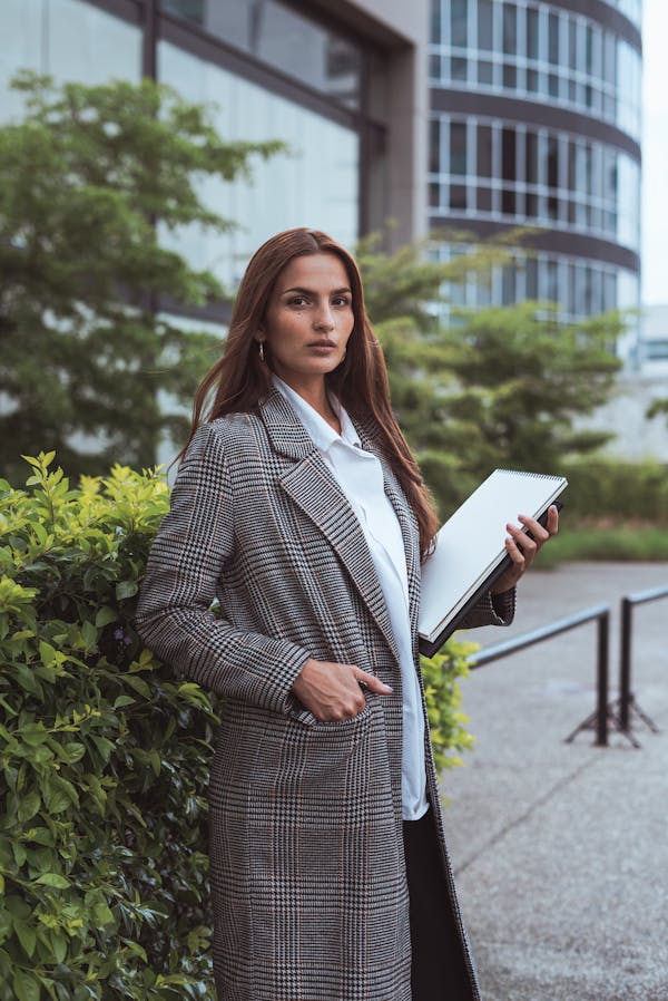 Confident woman in a stylish coat standing outside an office building holding a laptop.