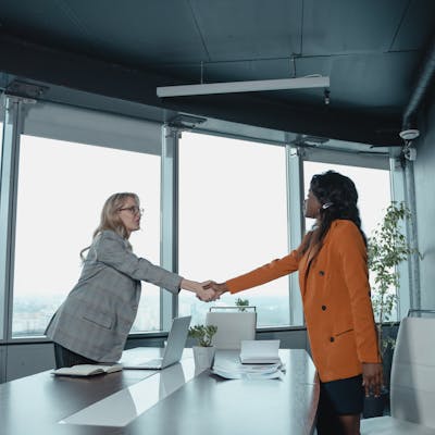 Two businesswomen shake hands across a conference table in a modern office setting.